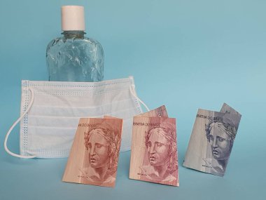 Brezilya banknotları, yüz maskesi, jel alkollü şişe ve mavi arka plan.
