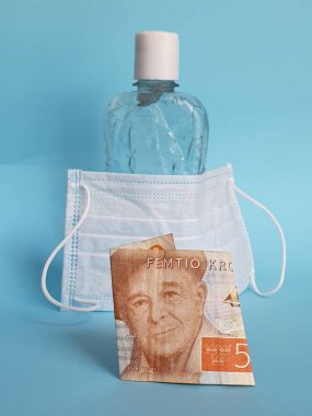 50 kronluk İsveç banknotu, yüz maskesi, jel alkollü şişe ve mavi arka plan.