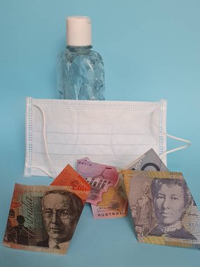 Avustralya banknotları, yüz maskesi, jel alkollü şişe ve mavi arka plan.