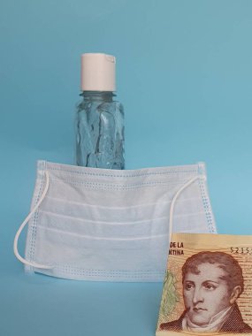 Arjantin banknotu 10 peso, yüz maskesi, jel alkollü şişe ve mavi arka plan.