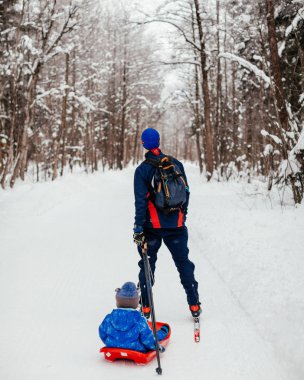 Kayak yapan baba, karlı bir ormanda kızakla bir çocuk taşıyor.