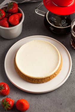 Yuvarlak tabakta hindistan cevizli cheesecake. Arka planda, bir çaydanlık, sade kahve ve bir kupa taze çilek. Gri arkaplanda yemek fotoğrafı.
