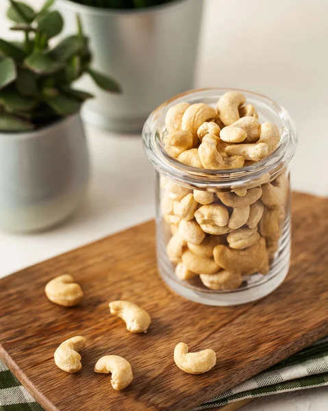 Veel cashewnoten in een glazen container op een houten plank op een lichte achtergrond. Op de achtergrond, groene planten in potten. — Stockfoto