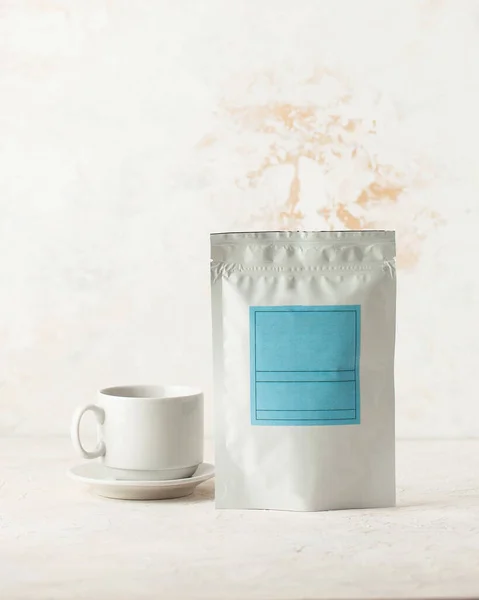 Aluminum bag for tea, coffee, seasonings and other bulk substances with a blue label for signature on a light background close-up. A tea bag and a white mug on a saucer.