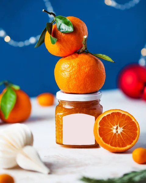 Citrus tangerine jam in a glass jar with a label for typing close-up on a festive Christmas background with Christmas tree toys. There\'s fresh citrus on the jar.