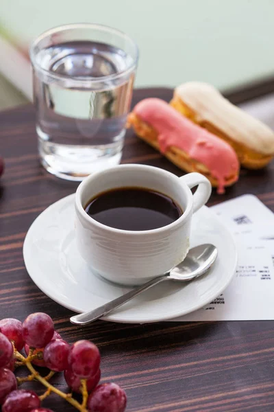 Белая кружка с черным кофе в центре, под блюдцем кофе, на заднем плане десерт с эклерами и стакан питьевой воды . — стоковое фото