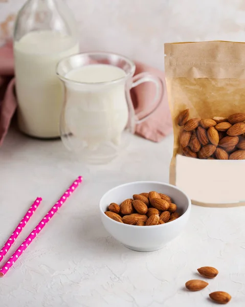A handful of almonds in close-up, next to a package of nuts with space for text and logo. In the background, a mug and a bottle of milk.