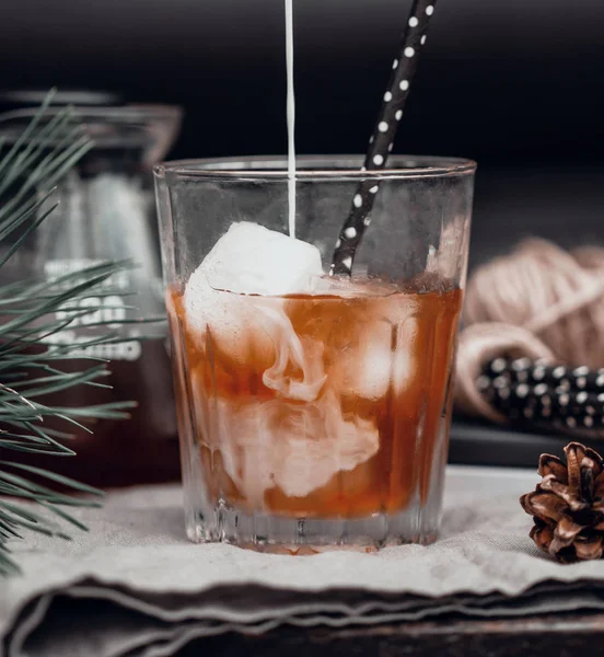 Göra kallt latte kaffe, en ränna mjölk hälls i svart kaffe i en transparent glas mugg på en mörk bakgrund. — Stockfoto