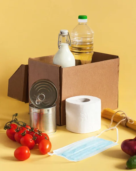 Safe delivery of food in a cardboard box with food supplies and disposable mask and gloves for the period of quarantine isolation on a yellow background. Food delivery, donation, contactless food delivery.