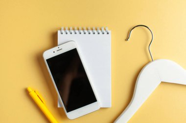  İnternetten alışveriş konsepti. Beyaz defter, kalem, cep telefonu ve sarı arka planda elbise askısı. Flatlay. Boşluğu kopyala. 