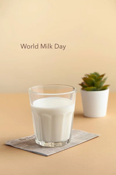 A mug of milk and a green plant in a pot on a brown background. Lettering Milk Day. June 1st. Copy space. Vertical food photo.