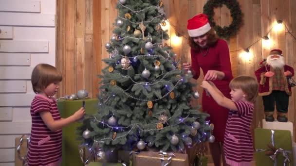 Мама помогает своим маленьким дочерям украсить елку перед Новым годом, деревянный фон — стоковое видео