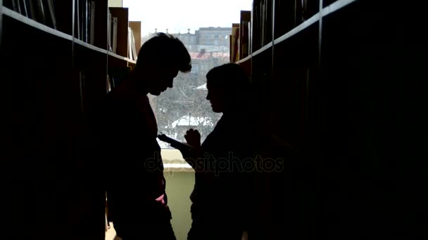 Estudante universitário de silhueta conversa entre as estantes de uma biblioteca — Vídeo de Stock