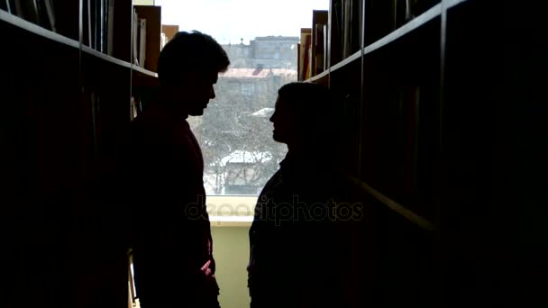 Estudante universitário silhueta conversa entre as estantes de uma biblioteca, companheiro sorridente. de perto — Vídeo de Stock