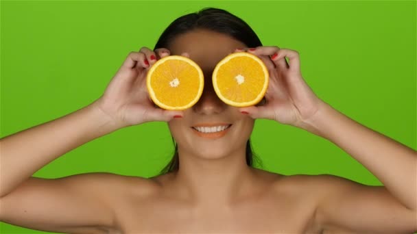 Девушка с красивым ярким макияжем покрывает глаза половинками апельсинов. Зеленый экран — стоковое видео