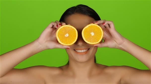 Девушка с красивым ярким макияжем покрывает глаза половинками апельсинов. Зеленый экран. Медленное движение — стоковое видео