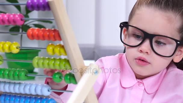 Маленькая девочка в очках сидит за столом и говорит разноцветным счетам. Закрыть — стоковое видео