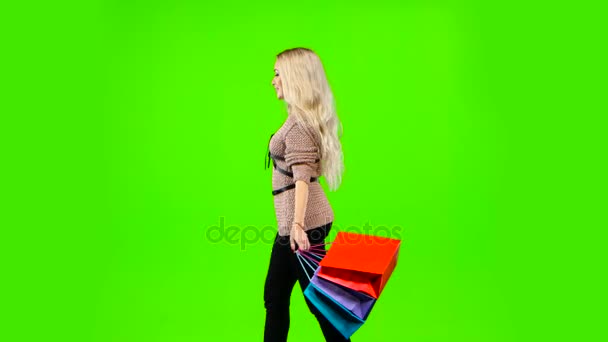 A szőke lány sok különböző színű csomagot hordoz. Zöld vászon. Oldalnézet — Stock videók