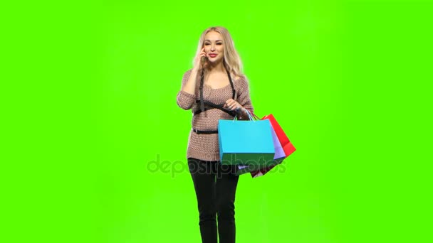 Menina loira carrega um multi pacotes coloridos, falando ao telefone e sorri docemente. Tela verde — Vídeo de Stock