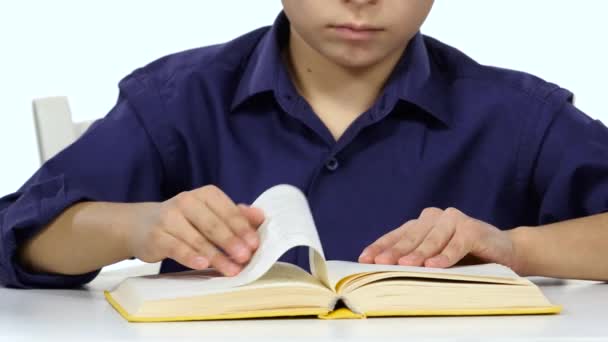 Мальчик сидит за столом и листает страницы книги. Белый фон. Закрыть — стоковое видео
