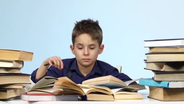 Leepy kisfiú ül az asztalnál, és fáradtan leafing keresztül egy könyvet. Kék háttér. — Stock videók