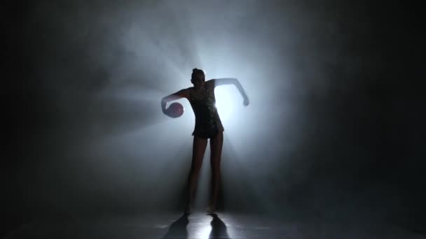 Gymnaste avec la balle dans ses mains faisant des mouvements acrobatiques. Fond noir. Arrière léger. Silhouette — Video