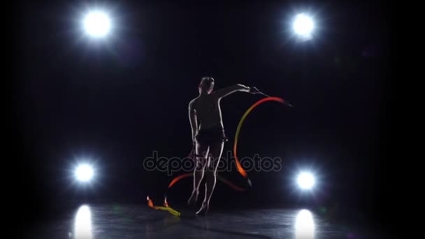 Gimnasta con la cinta en las manos haciendo movimientos acrobáticos. Fondo negro. Luz trasera. Movimiento lento — Vídeo de stock