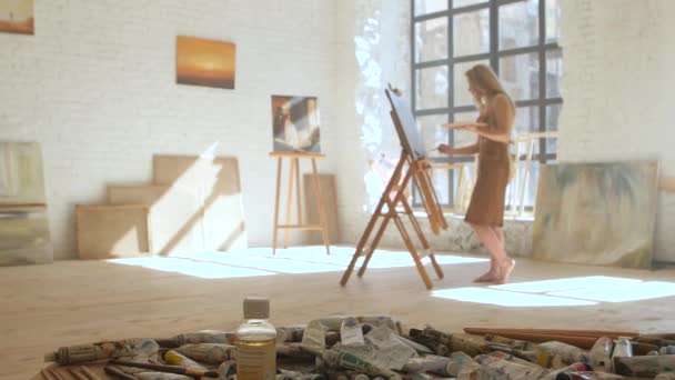 Chica artista pinta sobre lienzo sobre caballete y sostiene paleta en taller de arte — Vídeos de Stock