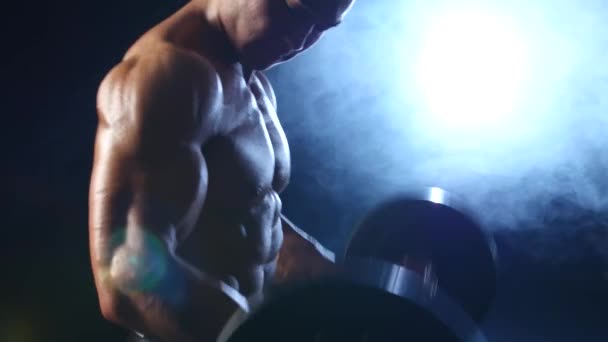 Bodybuilder levanta una barra para entrenar bíceps. Fondo de humo negro — Vídeos de Stock