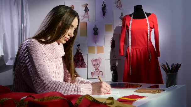 Créateur de mode dessine un croquis d'une robe rouge, à l'extérieur de la fenêtre déjà soir — Video