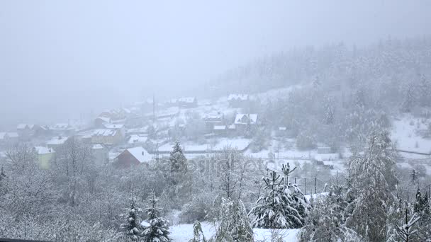 Nieve cae en pequeño pueblo a los pies de la colina arbolada — Vídeos de Stock