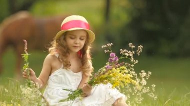 Bebek sarafan ve şapka glade oturur, kır çiçekleri ve gülümsüyor tutar. Ağır çekim
