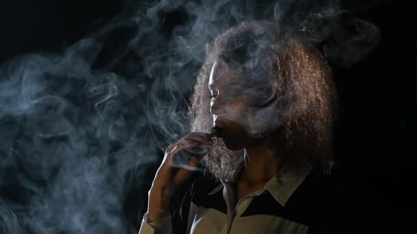 A afro-americana fuma um cigarro electrónico e deixa sair fumo da boca. Fundo preto. Movimento lento — Vídeo de Stock