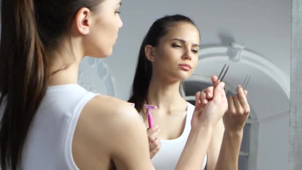 Chica sostiene un par de pinzas y una navaja de afeitar, y elige qué hacer con la depilación — Vídeo de stock