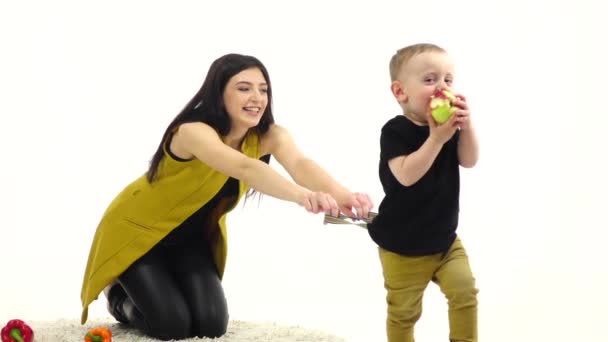 Menina senta-se no chão, ao lado dos vegetais mentira, ela tenta segurar o bebê. Fundo branco. Movimento lento — Vídeo de Stock