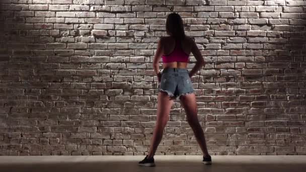 Жінка сексуальна у верхній частині врожаю і джинсові шорти танцюють тверк на цегляній стіні. Повільний рух — стокове відео