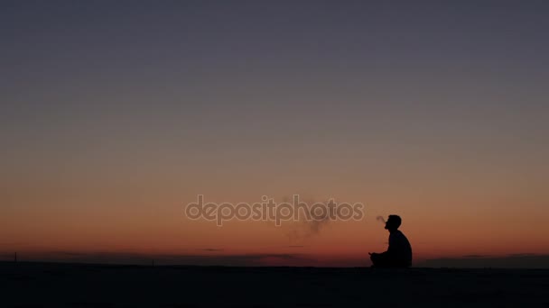 Srác ül a homokba, és dohányzik egy elektronikus cigaretta ellen a naplementét. Sziluettjét — Stock videók