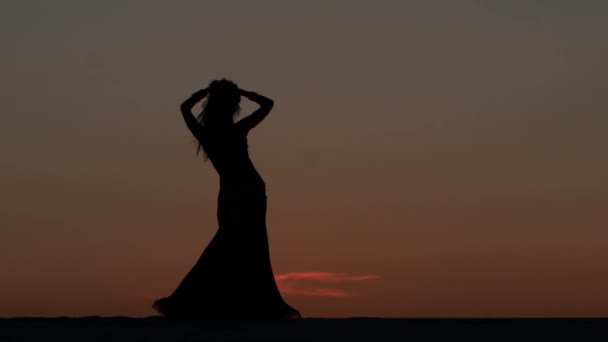 Menina está dançando barriga dançando contra o belo pôr do sol na praia. Silhuetas — Vídeo de Stock