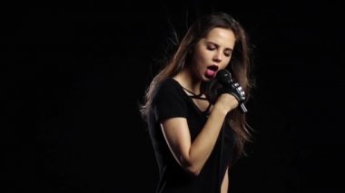 Şarkıcı enerjik şarkı söylüyor, saçları rüzgarda çırpınan. Siyah arka plan. Ağır çekim