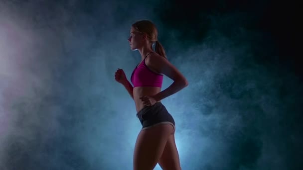 Sportovní fitness dívka běží na černém pozadí osvětlena reflektorů v kouři. Zpomalený pohyb. Detailní záběr — Stock video