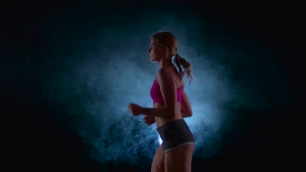 Chica corriendo sobre un fondo negro iluminado por el foco en el humo. En cámara lenta. De cerca. — Vídeos de Stock