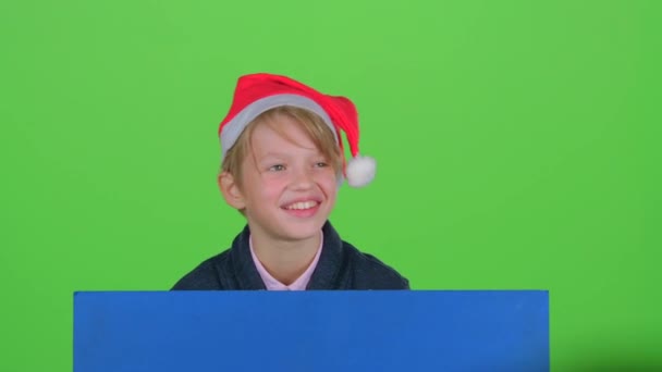 Niño en sombrero de Navidad aparece periódicamente escondido detrás de un tablero en una pantalla verde — Vídeos de Stock