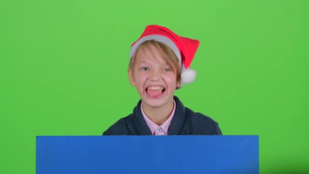 Niño apareció por detrás de un cartel azul muestra sus dientes sacude la cabeza y las curvas de caras y escondido de nuevo. Pantalla verde — Vídeos de Stock