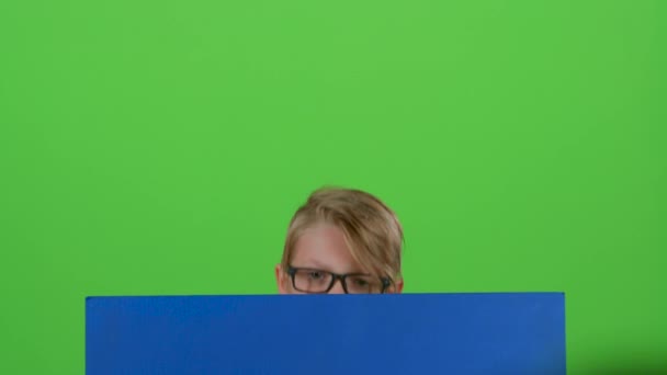 Chłopiec dziecko w okularach wyłania się zza deski i macha na zielonym ekranie — Wideo stockowe