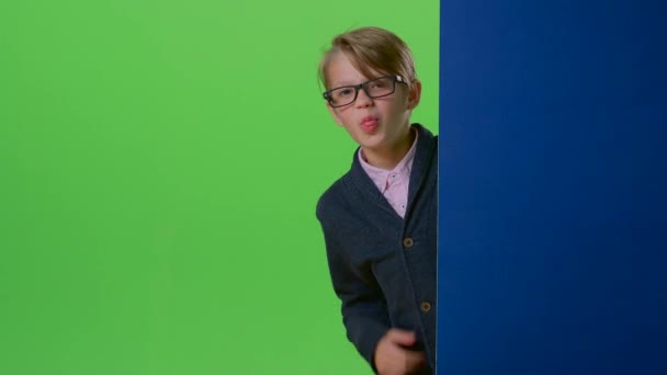 Teen in glasses comes from behind the wall and shows tongue on a green screen — Stock Video