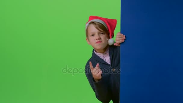 クリスマス帽子の 10 代の少年がボードと緑色の画面に彼の人差し指を振って後ろから現れる — ストック動画