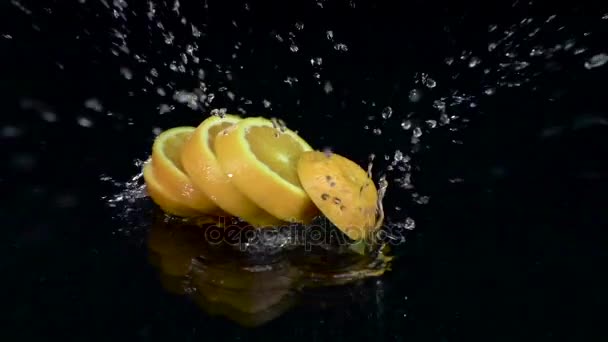 La naranja se disuelve en rodajas cuando cae al agua. Fondo negro. Movimiento lento — Vídeo de stock