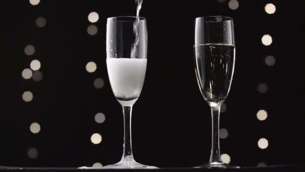 Шампанское наливают в два прозрачных бокала. Бокех мигает на чёрном фоне — стоковое видео
