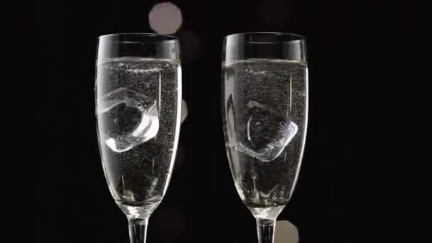 Twee glazen van champagne gevuld met een ice cube in elk. Bokeh Blinkende zwarte achtergrond. Close-up — Stockvideo