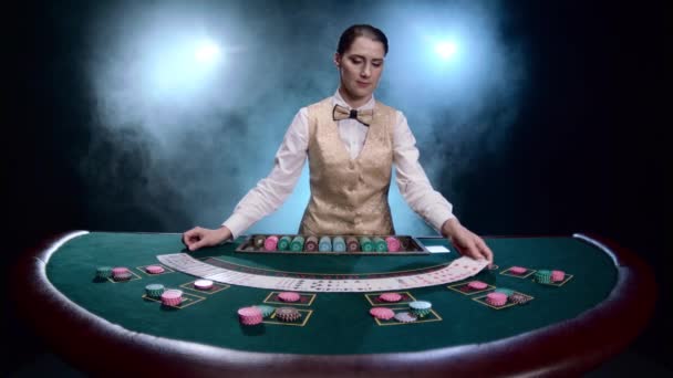 Mädchen Croupier in einem Casino mischt Karten auf schwarzem, rauchigen Hintergrund mit Scheinwerfern. Zeitlupe — Stockvideo